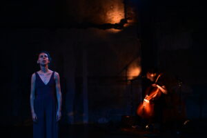 On voit deux femmes. L'une se tient debout, l'autre en fond de scène, joue du violoncelle.
